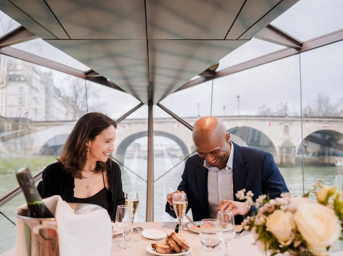 Coppia sorridente a cena con calici di champagne sotto un tetto di vetro, ponte fuori, tour in barca a Parigi