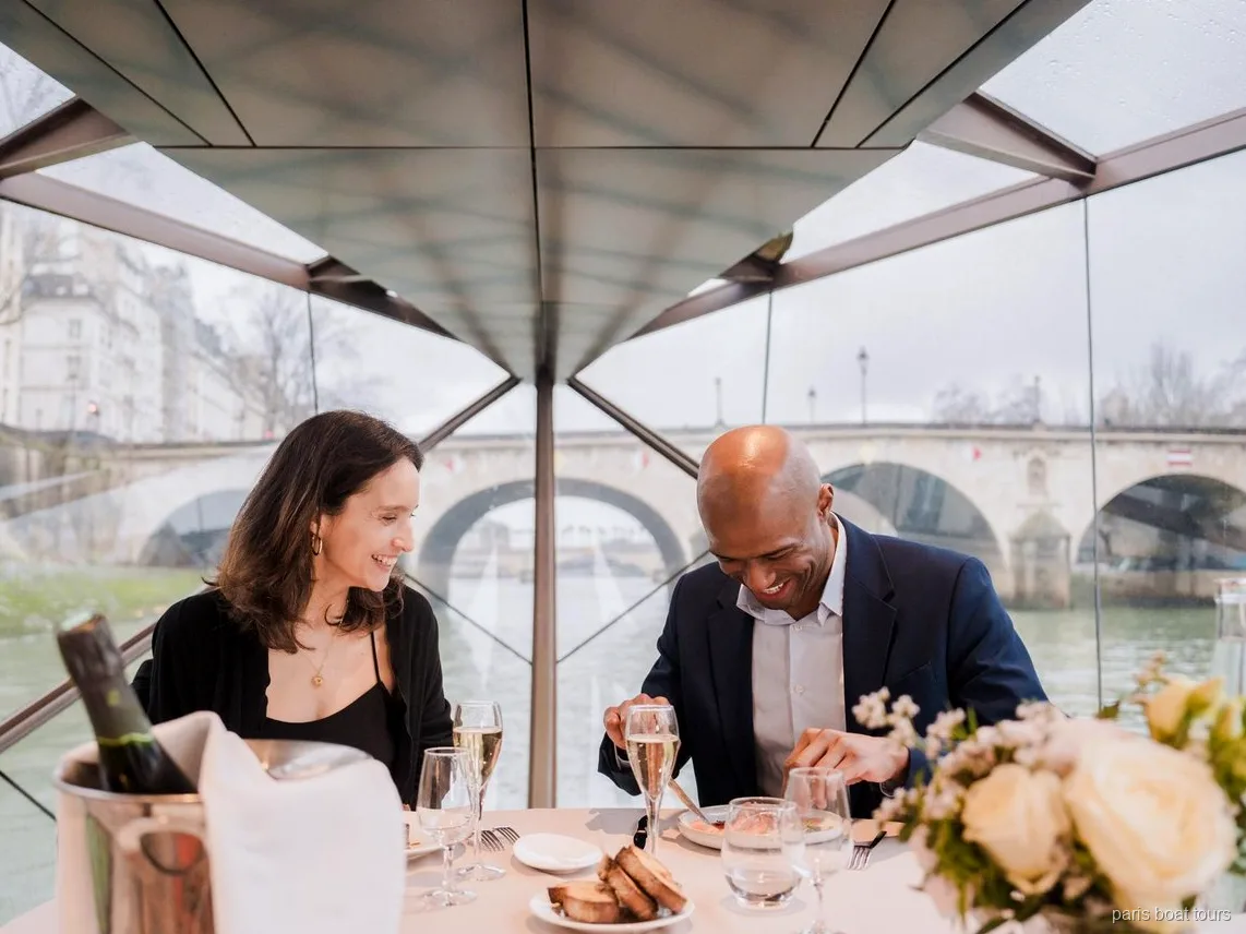 Couple smiling over dinner with champagne glasses under a glass roof, bridge outside, on Paris boat tours
