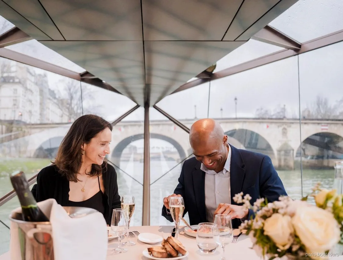 Par som ler under middagen med champagneglas under ett glastak, bro utanför, båtturer i Paris