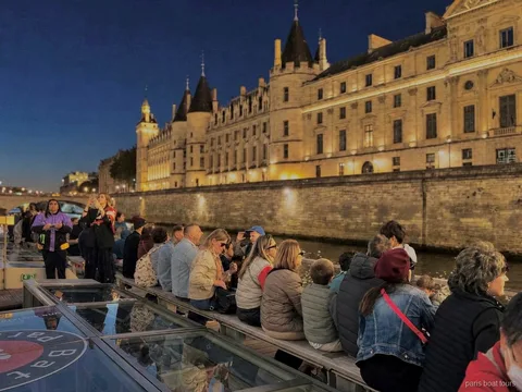 Fahrgäste nachts an Deck vor einem angestrahlten historischen Gebäude am Ufer, Bootstouren in Paris