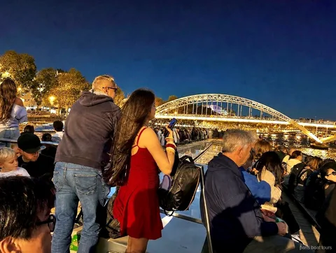Fahrgäste fotografieren nachts von Deck eine beleuchtete Bogenbrücke für Fußgänger, Bootstouren in Paris