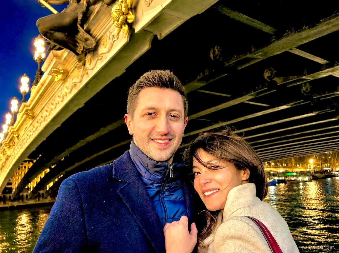Lächelndes Paar unter einer verzierten beleuchteten Brücke über der Seine bei Nacht, Bootstouren Paris