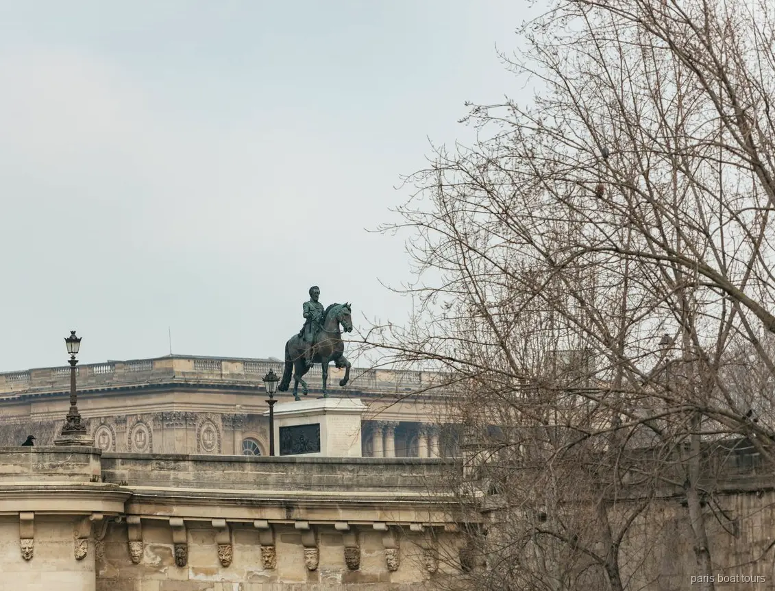 Estàtua eqüestre de bronze sobre un pont de pedra entre arbres nus, vista en passejades en vaixell per París