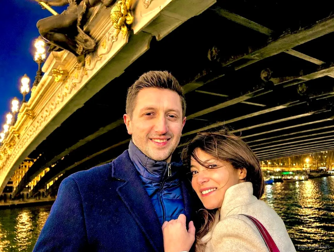 Parella somrient sota un pont ornamentat i il·luminat sobre el Sena de nit, passejades en vaixell per París