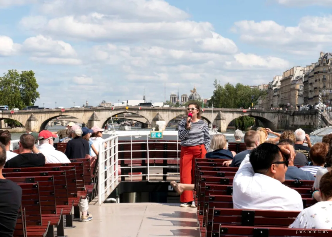 Guia amb micròfon parlant als passatgers a la coberta oberta d'un creuer per París vora un pont de pedra