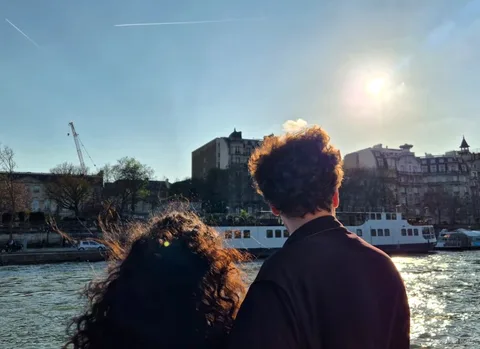 Dos passatgers d'esquena observen la riba del Sena i un vaixell amb el sol baix en un creuer per París
