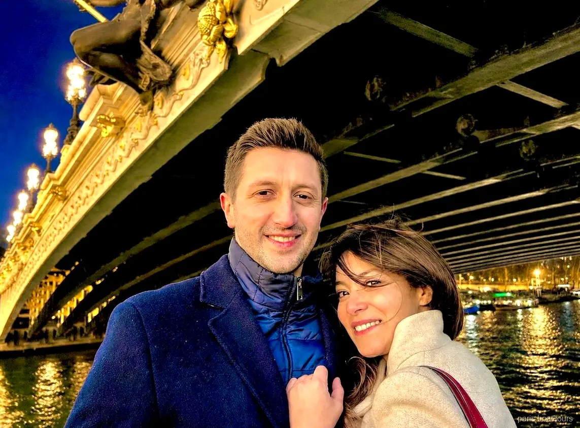 Coppia sorridente sotto un ponte ornato e illuminato sulla Senna di notte, giri in barca a Parigi