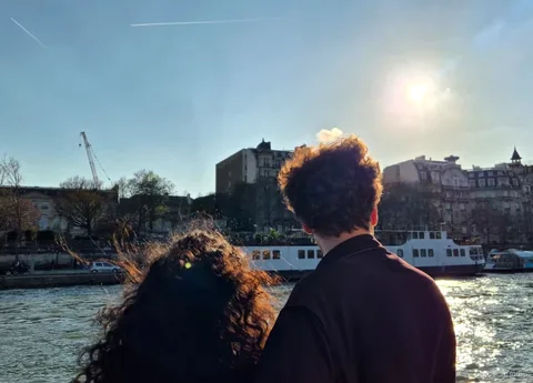 Due passeggeri di spalle guardano la riva della Senna e un battello con il sole basso in un tour in barca a Parigi