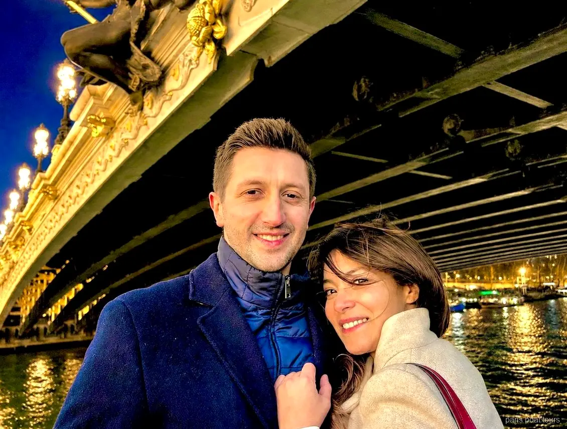 Couple souriant sous un pont orné et illuminé sur la Seine de nuit, croisières à Paris
