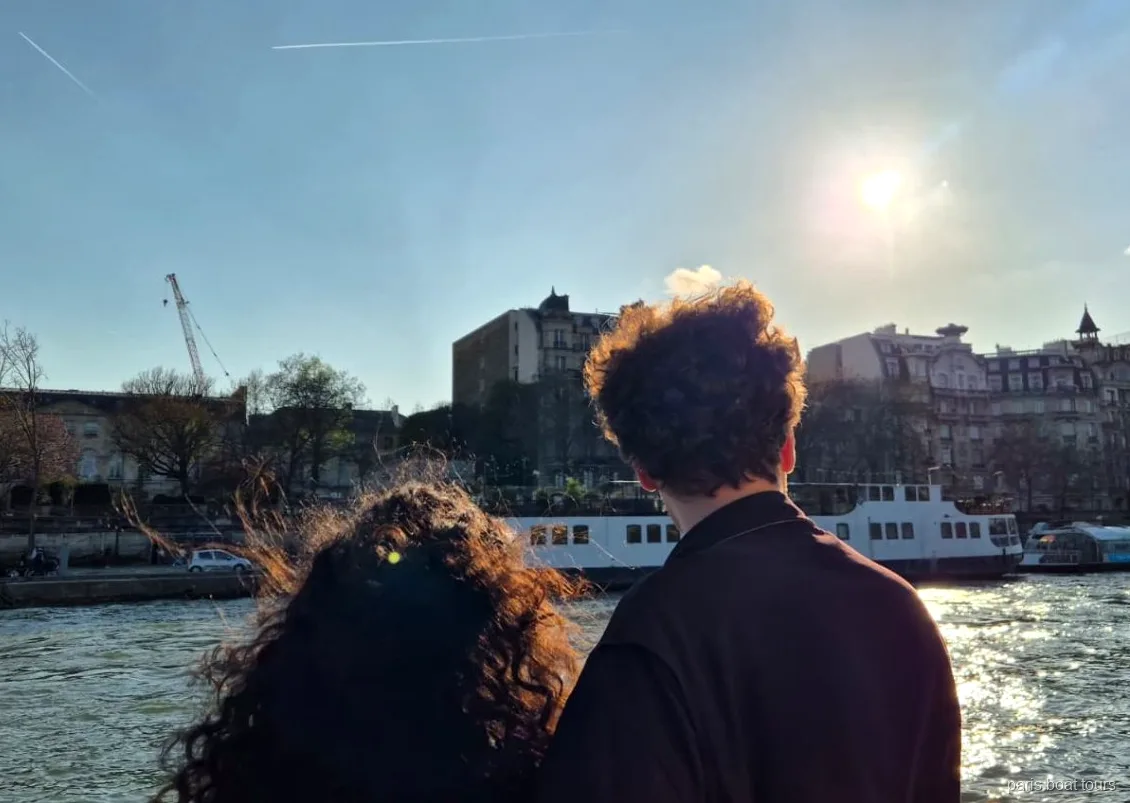 Dos pasajeros miran el Sena soleado y un barco amarrado desde la cubierta, paseos en barco por París