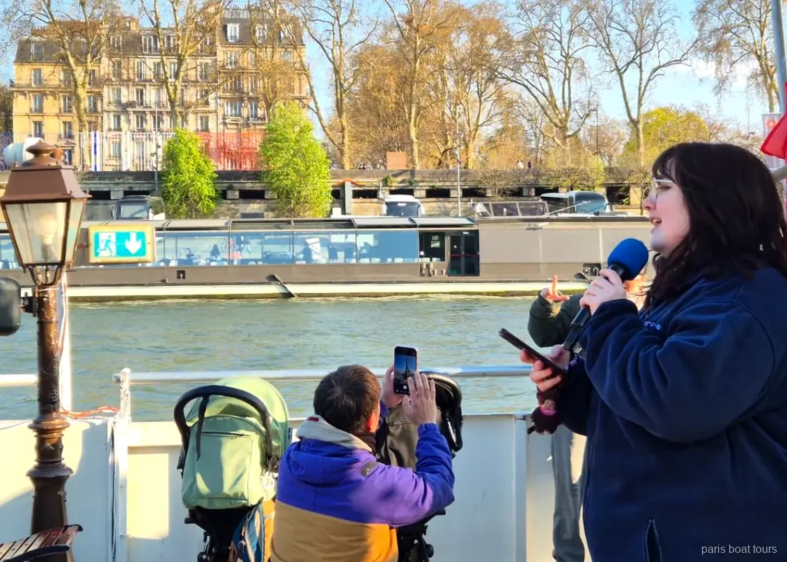 Guía hablando por micrófono en cubierta mientras los pasajeros graban el río, paseos en barco por París