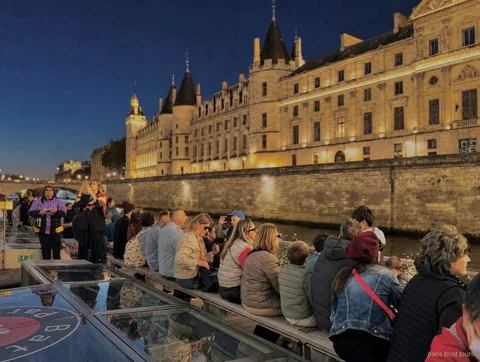 Pasajeros en cubierta de noche ante un edificio histórico iluminado en la orilla, paseos en barco por París