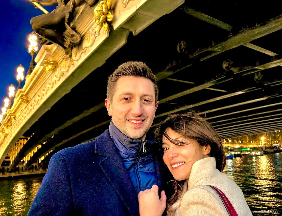 Pareja sonriente bajo un puente ornamentado e iluminado sobre el Sena de noche, paseos en barco por París