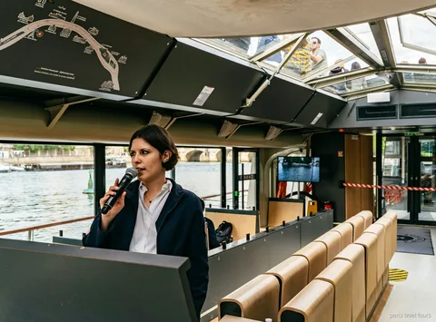 Guía con micrófono dentro de un barco con techo de cristal junto a un mapa de la ruta, paseos en barco por París