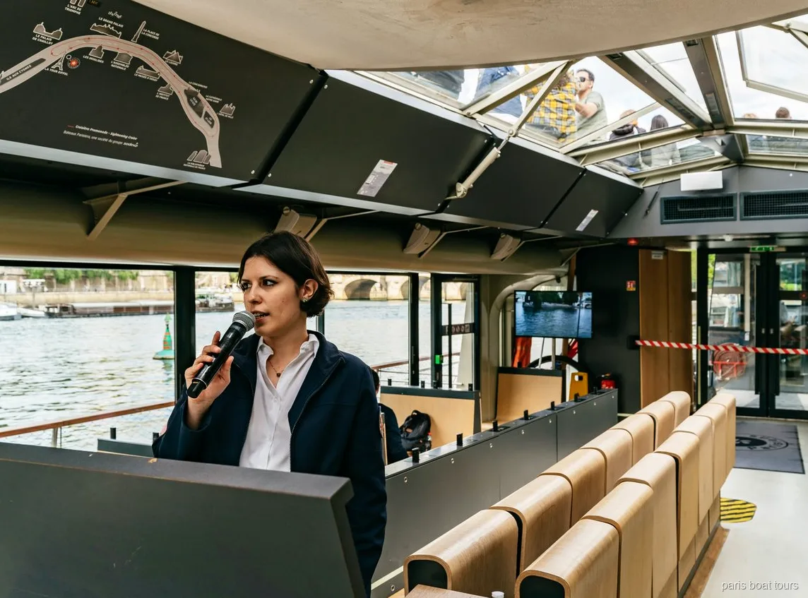 Guía con micrófono dentro de un barco con techo de cristal junto a un mapa de la ruta, paseos en barco por París