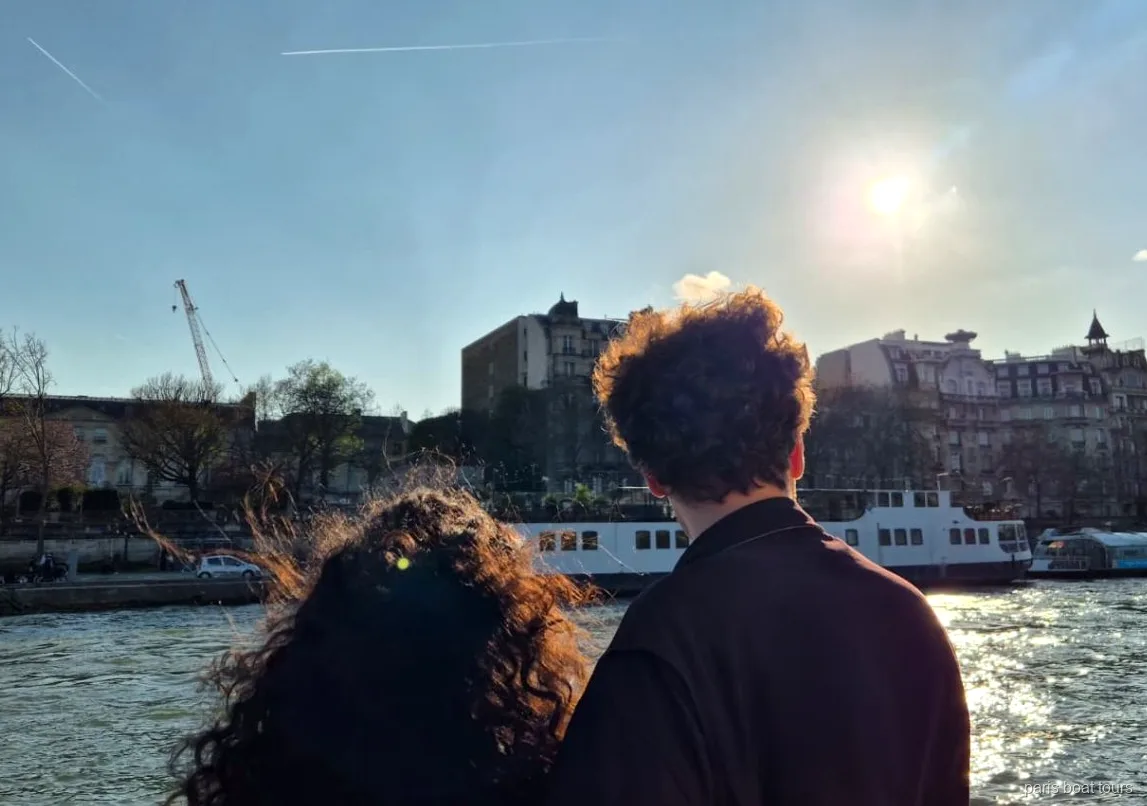Dois passageiros de costas olham a margem do Sena e outro barco sob o sol baixo em um passeio de barco em Paris