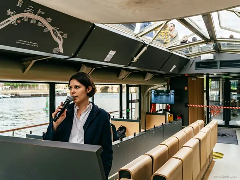 Guia com microfone dentro de um barco com teto de vidro ao lado de um mapa do trajeto, passeios de barco em Paris