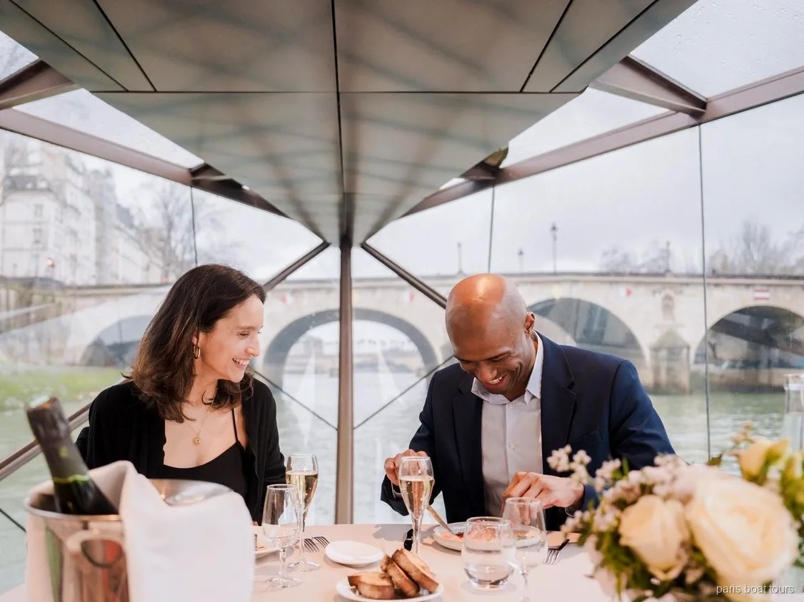 Couple souriant pendant le dîner avec des coupes de champagne sous un toit vitré, pont au dehors, croisières à Paris