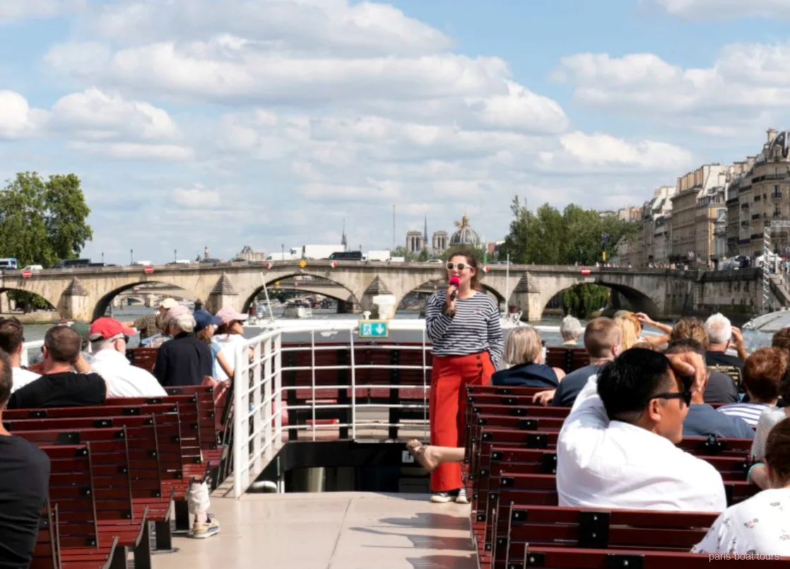 Guide med mikrofon talar till passagerare på öppet däck under en båttur i Paris vid en stenbro