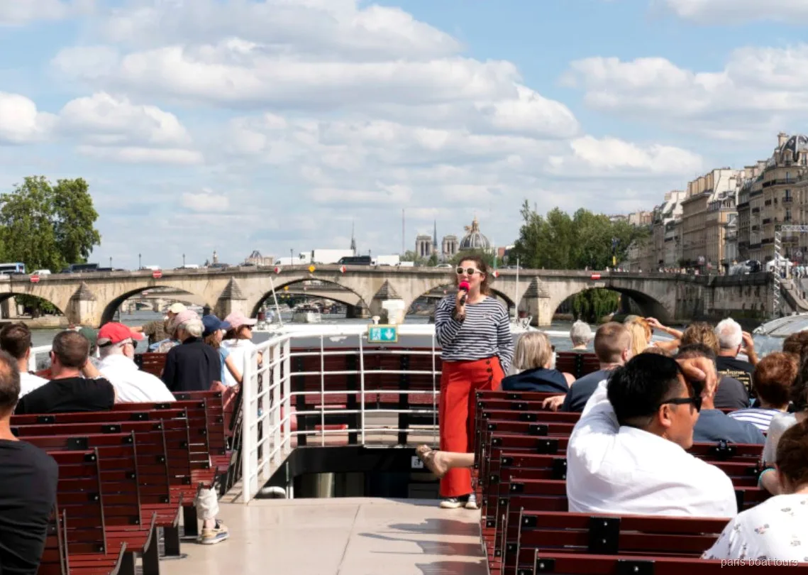 Guide with a microphone speaking to passengers on the open deck of a Paris boat tour beside a stone bridge