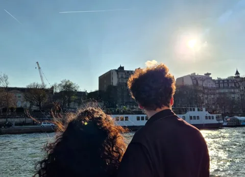 Zwei Passagiere blicken von Deck auf die sonnige Seine und ein vertäutes Schiff, Bootstouren in Paris