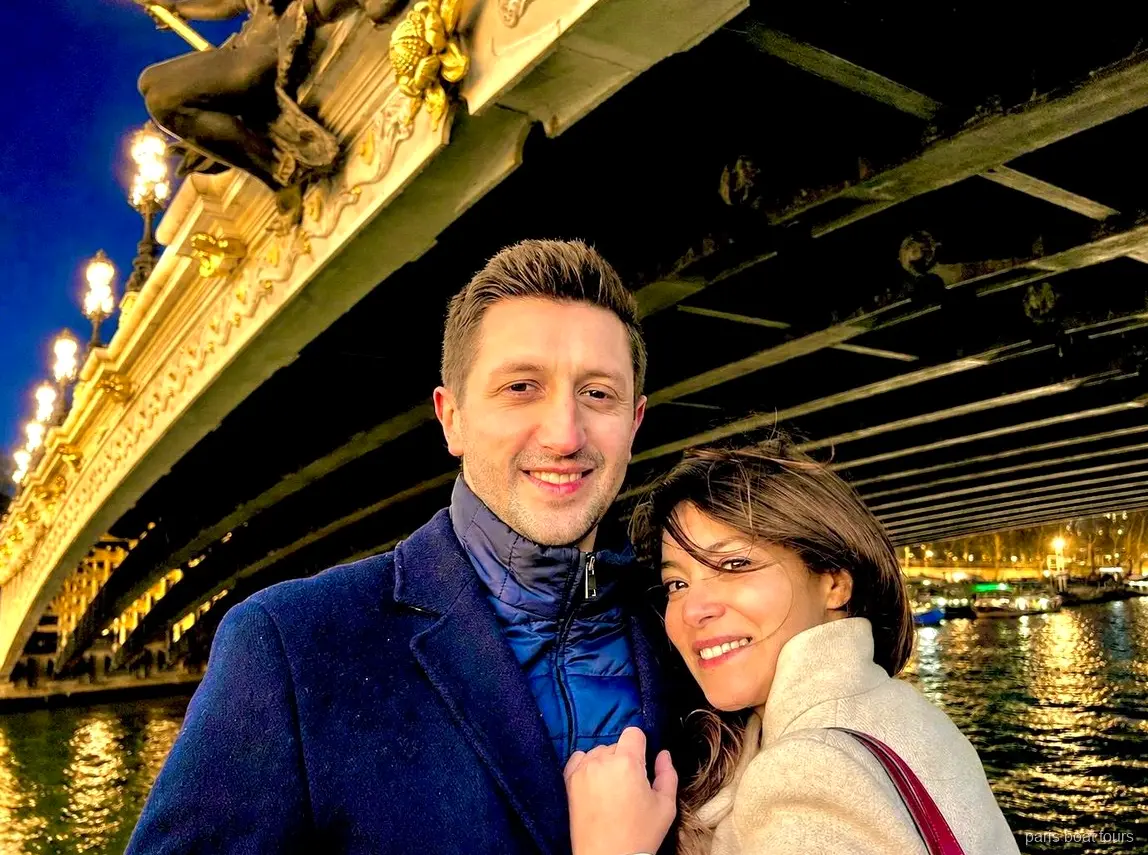 Smilende par under en udsmykket oplyst bro over Seinen om natten, bådture i Paris