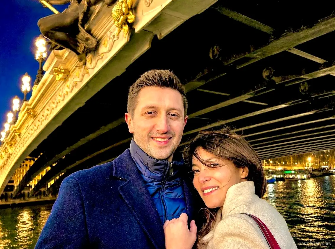 Casal sorridente sob uma ponte ornamentada e iluminada sobre o Sena à noite, passeios de barco em Paris