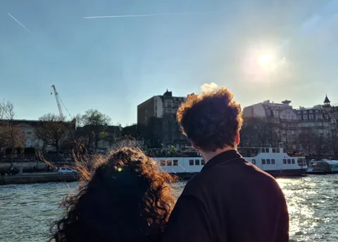 İki yolcu güvertede güneşli Seine nehrine ve demirli bir tekneye bakıyor, Paris tekne turları