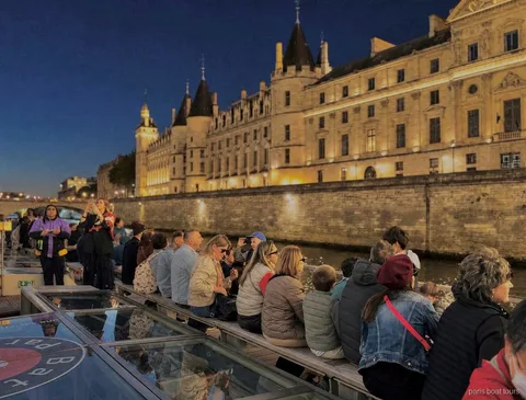 Passengers on deck at night facing a floodlit historic building on the bank, Paris boat tours