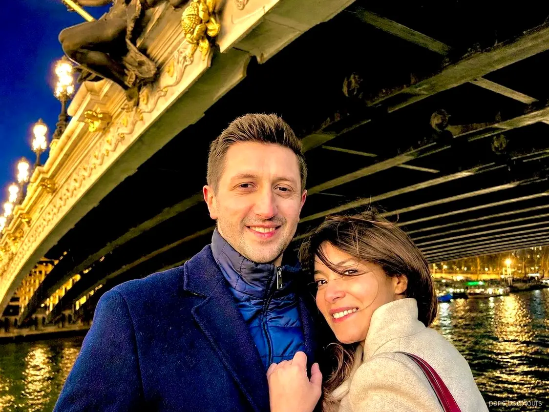 Smiling couple posing beneath an ornate lit bridge over the Seine at night, Paris boat tours