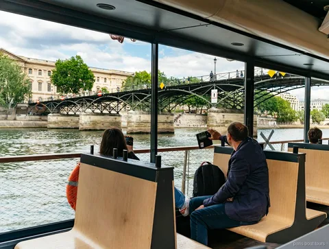 Passagerare på träbänkar inne i båten tittar på en gångbro över Seine, båtturer i Paris