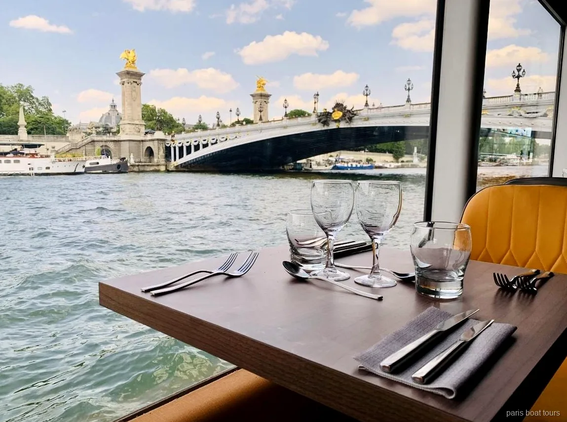 paris boat tours のディナークルーズ船で、窓際に食器とグラスが並ぶテーブル席とセーヌ川に架かるアレクサンドル3世橋の眺め