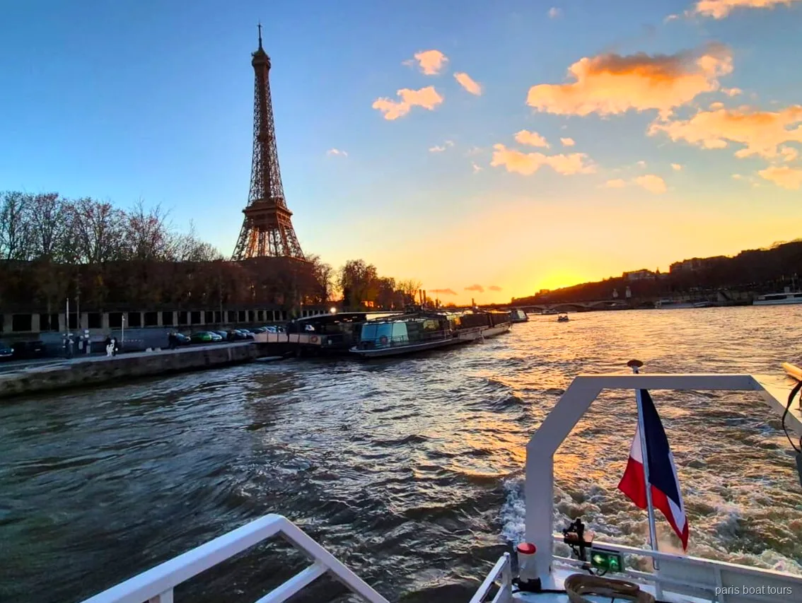 夕焼け空にそびえるエッフェル塔を、フランス国旗がはためくパリのボートツアーの船尾から望む様子、航跡の向こうにセーヌ川が広がる