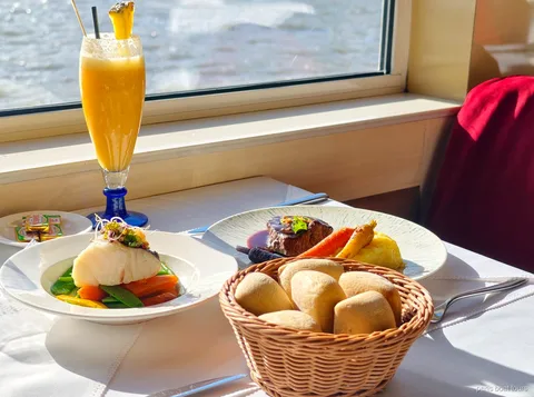 Taula vora la finestra amb peix i carn de vedella, cistella de pa i un suc de fruita, vista al riu, creuers per París