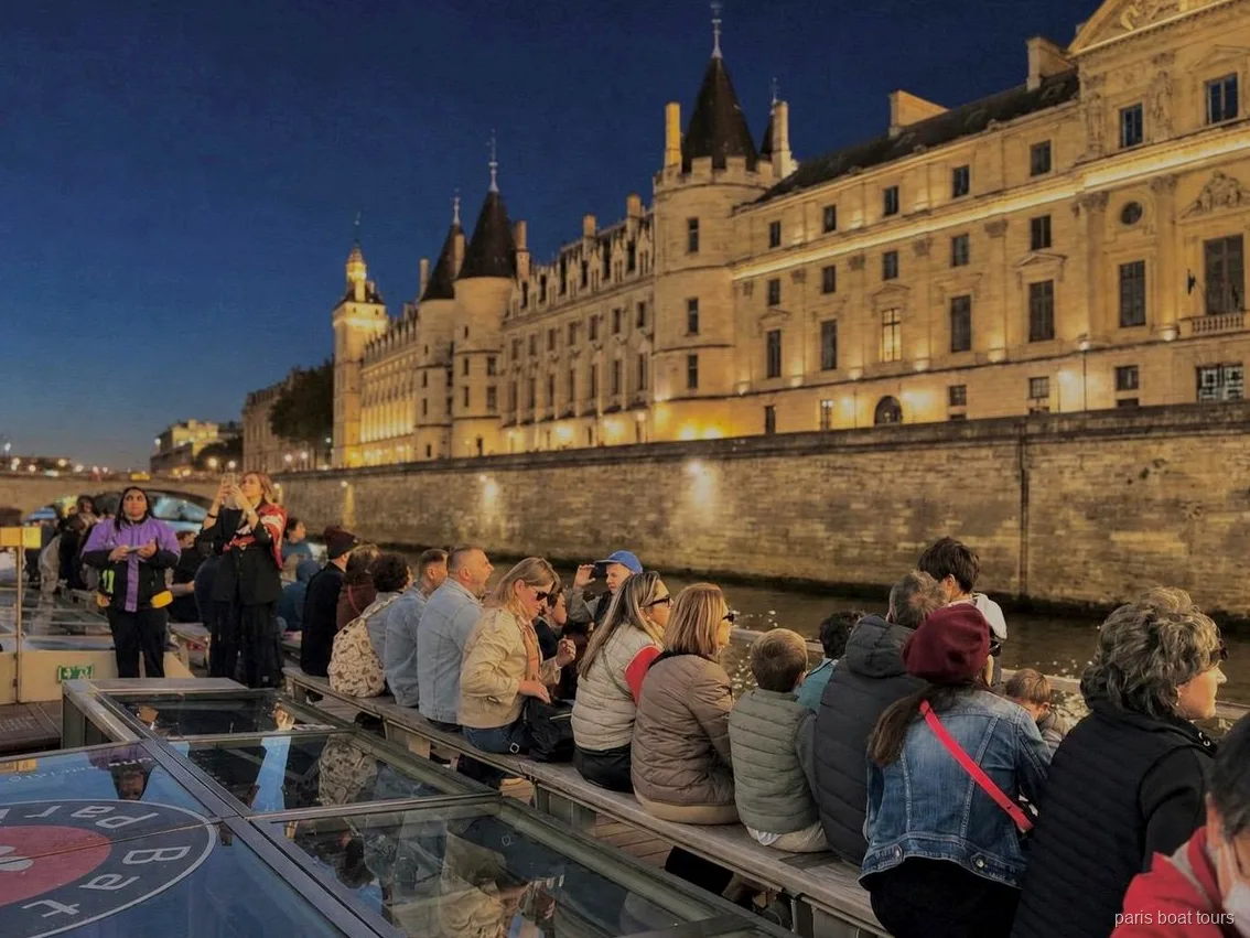 Passagerare på däck på kvällen framför en upplyst historisk byggnad vid stranden, båtturer i Paris