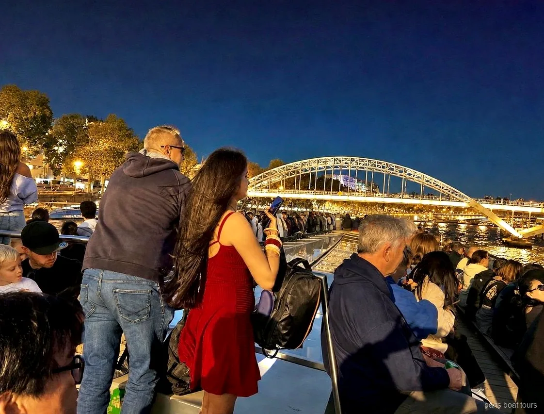 Passagerare fotograferar en upplyst bågformad gångbro från däck på natten, båtturer i Paris