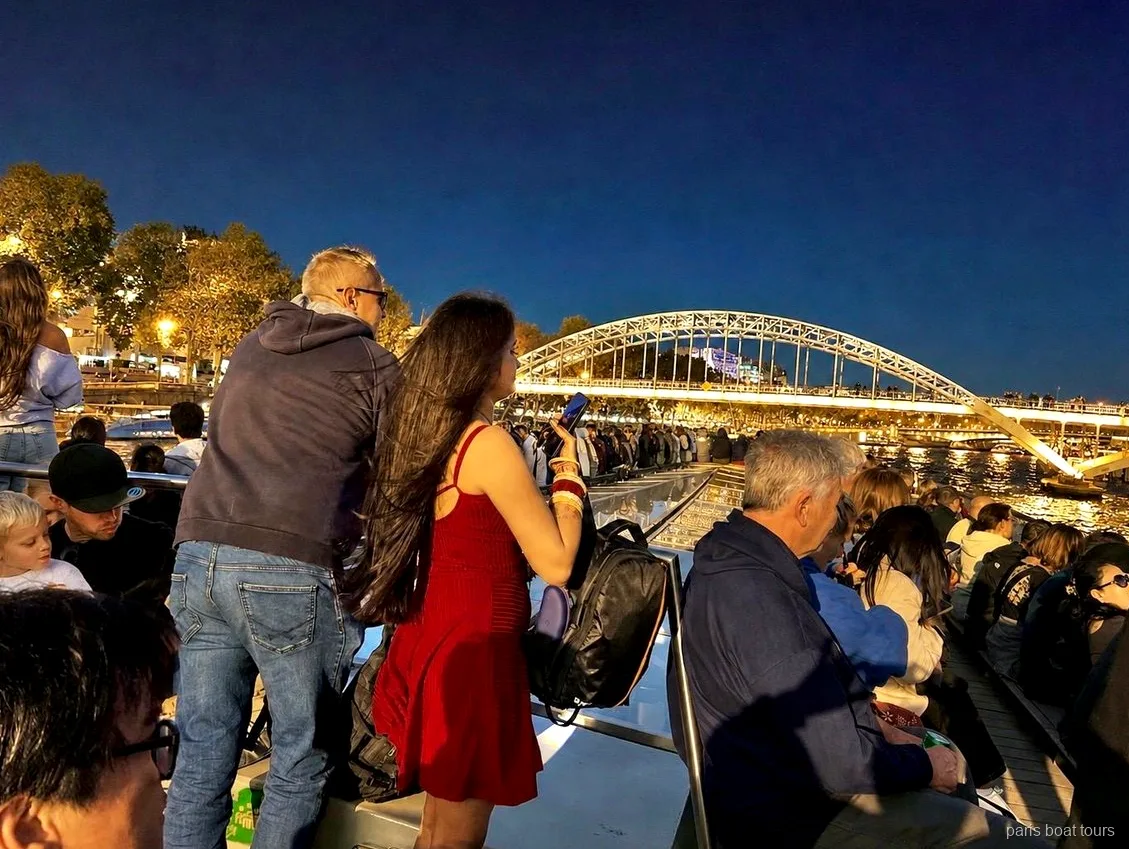 Passagiers fotograferen 's avonds vanaf het dek een verlichte voetgangersbrug met boog, boottochten Parijs